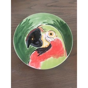 May include: A round, hand-painted ceramic plate featuring a parrot design. The parrot has a black beak, red chest, and yellow eye, set against a green background. The plate is a decorative item.