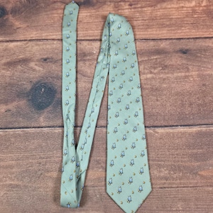 May include: A light sage green necktie featuring a repeating pattern of small, stylized praying hands in blue and white, interspersed with tiny gold-colored circular accents. The tie has a pointed tip and a smooth, woven fabric texture.