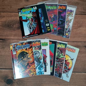 Pode incluir: Uma coleção de quadrinhos Spawn, cada um em uma capa protetora. As capas apresentam ilustrações dinâmicas com cores vibrantes, incluindo vermelho, preto e amarelo. Os títulos e as ilustrações são proeminentes.