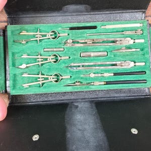 Peut inclure: Un ensemble d'instruments de dessin technique vintage présenté dans un étui noir avec un intérieur en velours vert. La collection comprend divers compas en métal, des diviseurs, des tire-lignes et des rallonges, rangés dans des compartiments.