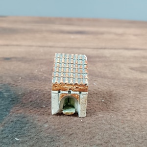 May include: A miniature, hand-painted model of a small building with a tiled roof. The structure has a light-colored facade with an arched opening and a detailed, textured roof in shades of brown and orange.