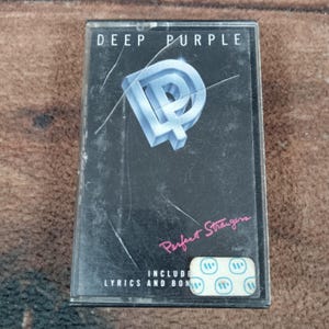 May include: A vintage cassette tape for the Deep Purple album "Perfect Strangers." The cover is black with the band's logo in a 3D-style design. The album title is written in pink script. Includes lyrics and bonus tracks.