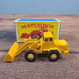 以下が含まれることがあります： 黒いタイヤと黄色のスコップが付いた、黄色のMatchbox Seriesのおもちゃのフロントエンドローダー。おもちゃは、Matchboxのロゴと赤いローダーのイラストが描かれたオリジナルの箱の前にあります。番号69が表示されています。