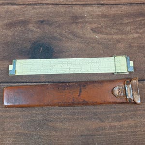 May include: An ivory-colored slide rule with black markings and a brown leather case. The slide rule has metal end pieces and a clear plastic cursor. The case is rectangular with a buckle closure.