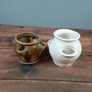 May include: Two ceramic strawberry pots. One is brown with a matching saucer, the other is white. Both pots have three openings for planting strawberries and small handles on the sides.