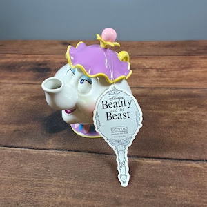 Könnte beinhalten: Eine Keramik-Teekanne in Form einer Figur mit cremefarbenem Gesicht, blauen Augen und rosa Mund. Sie hat einen violetten, gerüschten Deckel mit rosa Knauf. Ein weißes Etikett trägt die Aufschrift Disney's Beauty and the Beast, Schmid, Randolph, MA.