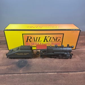 Puede incluir: Una locomotora de vapor a escala de color negro con techo de cabina rojo y un ténder a juego con la inscripción Pennsylvania-Reading Seashore Lines. El tren está colocado frente a una caja de cartón amarilla y naranja de Rail King by MTH Electric Trains.