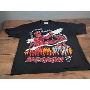 Puede incluir: Una camiseta negra de manga corta con un estampado gráfico de una figura de demonio roja con el texto THE MARK OF THE BEAST y DEMON. El diseño incluye llamas, una plataforma rocosa y un pequeño icono de demonio en tinta blanca, roja y naranja.