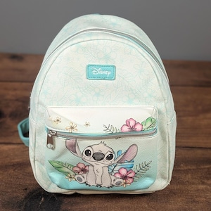 May include: A light blue and white mini backpack featuring a repeating tropical floral pattern. The front pocket displays a graphic of the character Stitch surrounded by pink hibiscus flowers and green leaves, with a Disney logo patch above.
