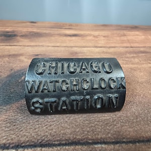 May include: A black, rectangular metal object with the embossed text "CHICAGO WATCHCLOCK STATION". The object has a curved top, possibly a vintage or antique item.