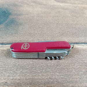 May include: A red multi-tool pocket knife with a silver blade and various tools. The knife features a gold emblem and a small metal loop for attaching to a keychain. The knife is closed, showing its red casing.