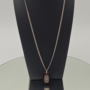May include: A silver-toned curb chain necklace featuring a rectangular pendant with rounded edges. The pendant has a dark, glossy border surrounding a central metallic inlay engraved with the capital letter M in a serif font style.