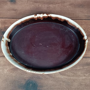 Puede incluir: Una fuente ovalada de cerámica con un interior burdeos oscuro. El borde tiene un diseño moteado blanco y marrón. El plato probablemente se utiliza para servir alimentos y tiene un aspecto rústico y hecho a mano.