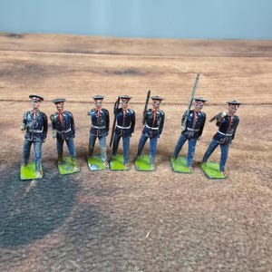 Puede incluir: Un conjunto de seis soldados de juguete antiguos. Cada soldado viste un uniforme azul oscuro con detalles rojos y pantalones azules. Están sobre bases verdes y sostienen armas. Las figuras están dispuestas sobre una superficie de madera.
