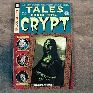 May include: A comic book titled "Tales From The Crypt" with a teal and red cover. The cover features the title in large white letters, the text "Terror" in red, and illustrations of ghouls. The comic is published by Papercutz.