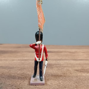 May include: A miniature toy soldier in a red jacket, black pants, and a tall black hat, holding a flag. The soldier is standing on a rectangular base with text. The flag is orange with a design. The soldier is saluting.