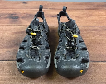 May include: A pair of dark gray closed-toe sandals featuring a protective rubber toe cap, black mesh panels, and a bungee cord lacing system with a yellow toggle. The sandals have a heel strap and a gray sole with a small yellow logo detail.