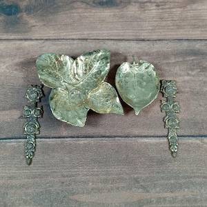 May include: A collection of decorative metal leaf-shaped dishes and floral-detailed serving pieces. The set includes a large three-leaf dish, a single leaf-shaped dish, and two ornate serving spoons, all in a metallic finish.