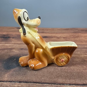 May include: A ceramic figurine of a tan dog with black ears and a white muzzle, depicted in a seated position. The dog is attached to a small, brown, two-wheeled cart with a hollow interior, featuring a pink center on the wheel hub.