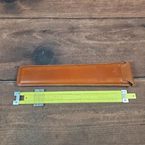 May include: A vintage slide rule with a yellow body and a metal frame, next to a brown leather case. The slide rule is a mathematical tool used for calculations. The case is rectangular and has a logo embossed on it.