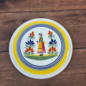 May include: A round, white ceramic plate with a yellow and blue border. The center features a folk art illustration of a woman in traditional dress, surrounded by floral designs. The plate is approximately 8 inches in diameter.