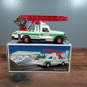 May include: A vintage Hess toy truck with a red extending ladder, white cab, and green accents. The truck sits on a blue box with a picture of a gas station and a similar truck. The truck has the word "GASOLINE" and "HESS" printed on it.