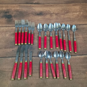 Puede incluir: Un juego de cubiertos vintage con mangos rojos y detalles dorados. El conjunto incluye tenedores, cuchillos, cucharas y cuchillos de mantequilla. Los utensilios están dispuestos sobre una superficie de madera.