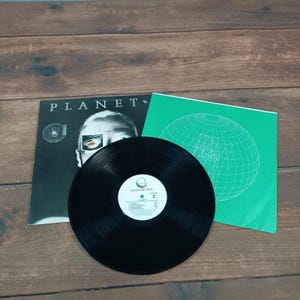 May include: A black vinyl record with a white label, resting on a wooden surface. The album cover features the word "PLANET=" and a graphic of a face. A green sleeve with a globe design is also visible.