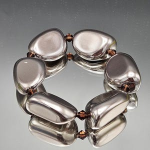 May include: A silver-toned bracelet featuring large, smooth, oval-shaped beads. The beads are connected by small, brown, faceted beads. The bracelet is displayed on a reflective surface.