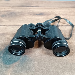 Könnte beinhalten: Schwarzes Fernglas mit weißem Rand um die Linsen und einem schwarzen Riemen. Das Fernglas hat eine strukturierte Oberfläche und ist vor einem Holzgrund platziert. Die Linsen sind klar und einsatzbereit.