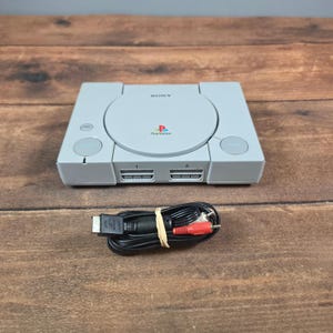 Puede incluir: Una consola de videojuegos Sony PlayStation de color gris claro con una tapa circular para discos que muestra el logotipo de PlayStation, dos puertos para mandos y un botón de encendido. Incluye un cable AV negro con conectores rojo y blanco, sujeto con una goma.