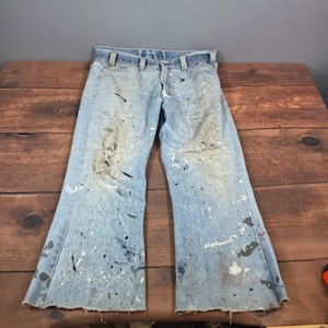 Puede incluir: Pantalones vaqueros de mezclilla azul claro con corte recto, cierre de botones y trabillas para cinturón. La tela muestra un desgaste intenso con numerosas salpicaduras de pintura en tonos blanco, negro y marrón, y bajos deshilachados.