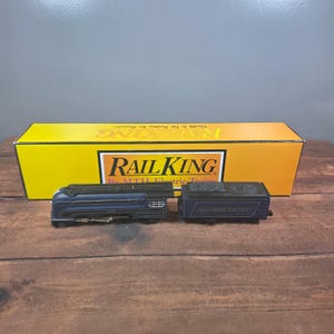 May include: A dark blue model steam locomotive and tender with silver accents, positioned in front of a yellow Rail King by MTH Electric Trains box. The locomotive features the text Baltimore and Ohio on the side of the coal tender car.