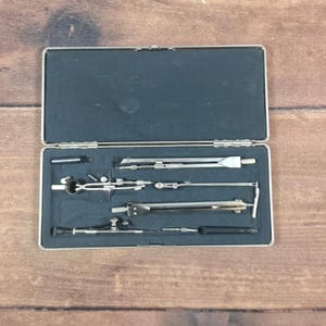 May include: A silver-toned metal drafting compass set housed in a rectangular case with a black foam interior. The set includes various precision drawing instruments, including dividers, a bow compass, and extension bars, all arranged in molded slots.
