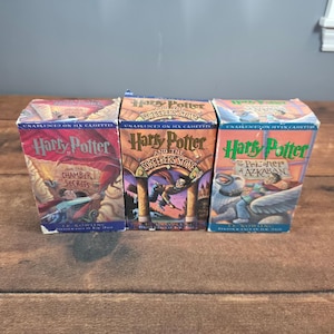 May include: Three vintage Harry Potter cassette tape boxes. The boxes are labeled "Harry Potter and the Chamber of Secrets", "Harry Potter and the Sorcerer's Stone", and "Harry Potter and the Prisoner of Azkaban". The boxes are on a wooden surface.