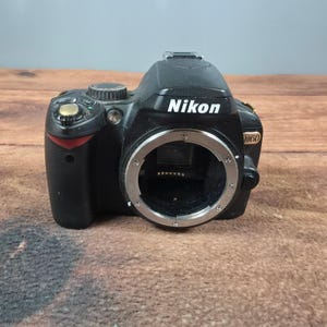 May include: A black Nikon D60 digital SLR camera body featuring a prominent silver lens mount, a gold-colored shutter release button, a mode dial, and the Nikon brand name printed in white on the front of the camera housing.