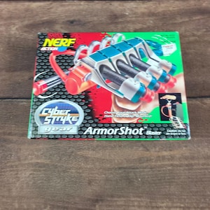 May include: A Nerf Action Cyber Stryke Gear ArmorShot Blaster toy box featuring a teal, white, and orange hand-mounted blaster design. The packaging displays the product name, a graphic of the blaster in use, and a red and black dotted background.