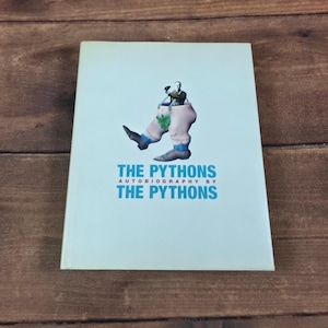 May include: A light blue book titled The Pythons Autobiography by The Pythons. The cover features a surreal illustration of a person's upper body emerging from a pair of large, pale legs wearing blue and brown shoes with a green leaf at the waist.