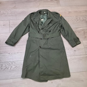 Puede incluir: Un abrigo tipo trench verde oliva con diseño de doble botonadura, cinturón en la cintura y puños abotonados. El abrigo tiene charreteras en los hombros y un cuello clásico. La longitud del abrigo es de aproximadamente 114 cm.