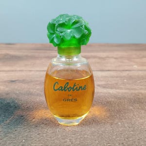 May include: A small, clear glass perfume bottle with a green, flower-shaped cap. The bottle contains a golden-colored liquid and features the word "Cabotine" in cursive script. The bottle is on a wooden surface.