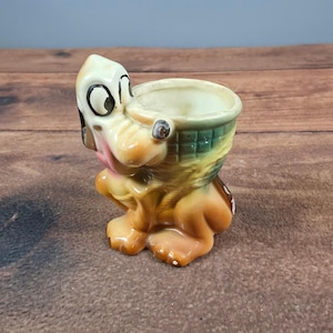 May include: A ceramic egg cup shaped like a cartoon dog with a tan body and a green basket on its back. The dog has large white eyes with black pupils, a pink tongue, and a textured basket weave pattern on the side of the container.