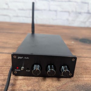 May include: A black YjHifi Audio amplifier featuring a vertical antenna, a power toggle switch with a red indicator light, and three circular control knobs on the front panel. The device has a rectangular metal housing and a power cord.