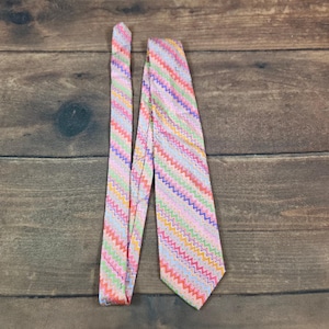 May include: A necktie featuring a repeating chevron or zigzag pattern in a vibrant color palette of pink, orange, light blue, purple, and green. The tie is constructed with a pointed tip and a narrow tail, displaying a consistent woven texture.