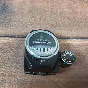 May include: A black Honeywell Pentax Meter with a silver dial and a black control knob. The meter has a circular dial with numbers and markings, including the text "HONEYWELL PENTAX METER". The control knob has numbers from 1 to 1000.