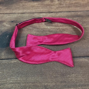 May include: A bright magenta bow tie featuring a subtle herringbone weave pattern. The bow tie includes an adjustable neck strap with a small black hardware slider and a dark fabric tag attached to the inner band near the adjustable mechanism.