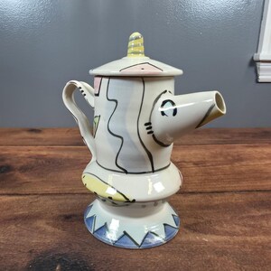 May include: A ceramic teapot with a lid, spout, and handle. The teapot is white with colorful abstract designs in yellow, pink, blue, and black. The base has a blue and white geometric pattern. The spout is angled and the handle is curved.