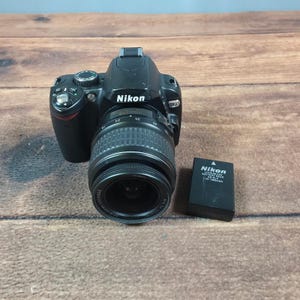 May include: A black Nikon D60 digital SLR camera with an attached 18-55mm lens and a separate black Nikon EN-EL9 lithium-ion battery pack. The camera body features a mode dial, shutter button, and the Nikon logo printed on the front housing.