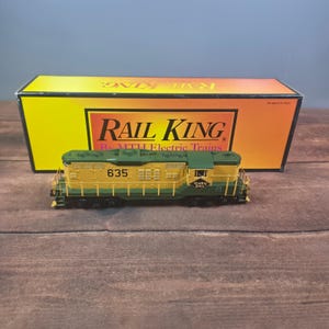 Può includere: Una locomotiva diesel in miniatura gialla e verde con il numero 635 e il marchio Reading Lines, posizionata davanti alla sua scatola di cartone rettangolare Rail King by MTH Electric Trains con un design a sfondo sfumato da giallo ad arancione.
