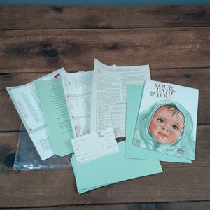 May include: A collection of vintage baby-themed ephemera, including a booklet titled "Your Baby & You" featuring a baby illustration. Other items include pale green envelopes, papers, and a clear plastic sleeve, all arranged on a wooden surface.