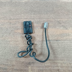 May include: A black electrical device with a coiled wire and a separate connector. The device has a rectangular shape with two red dots and a logo. The connector is silver and rectangular.